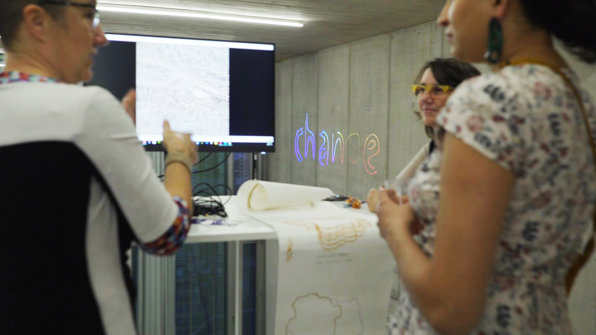 three artists stand together in a large concrete space. They are standing around a plotter with a very long sheaf of paper cascading out. In the background is a projection on the wall and a screen with art being projected.