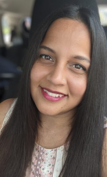 portrait headshot of Manal Siddiqui, long dark hair, smiling in front of a blurred background