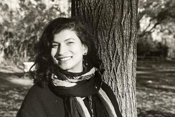 head shot of artist Prachi Khandekar - black and white, wearing a scarf and smiling while standing next to a tree outside