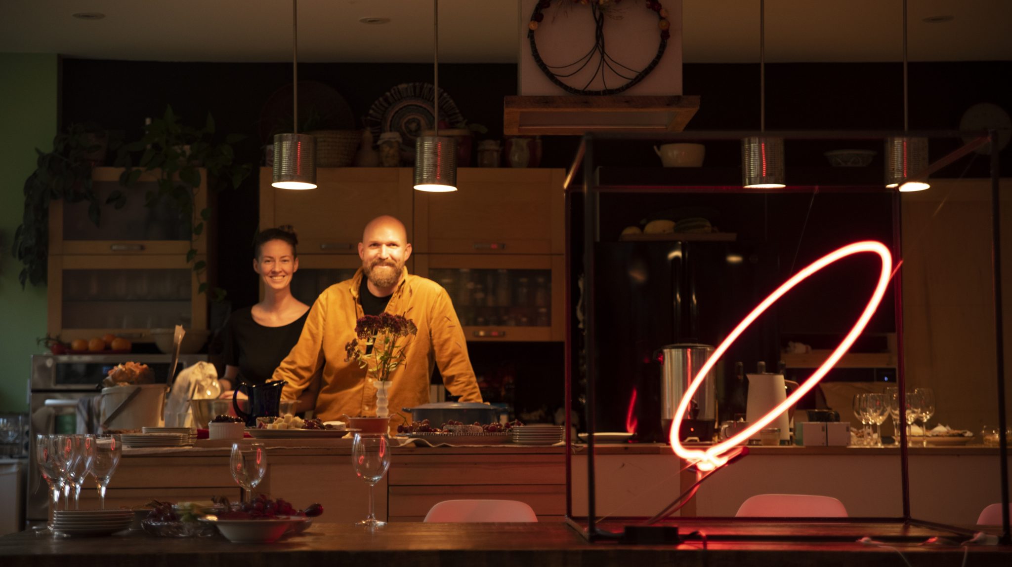 Mary and Bob smiling, looking at the camera. They are standing behind the kitchen counter with Hooria's neon installation, a red neon ring light, at the foreground